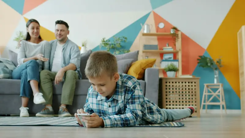Eltern sitzen auf dem Sofa, ihr Sohn liegt davor auf dem Teppich und spielt am Handy.