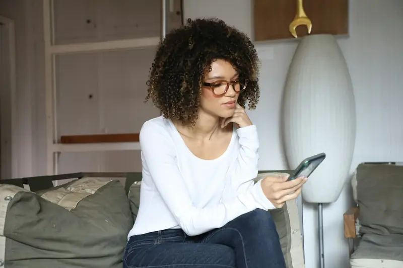 Eine Frau mit Brille sitzt auf dem Sofa und schaut besorgt auf ihr Telefon.
