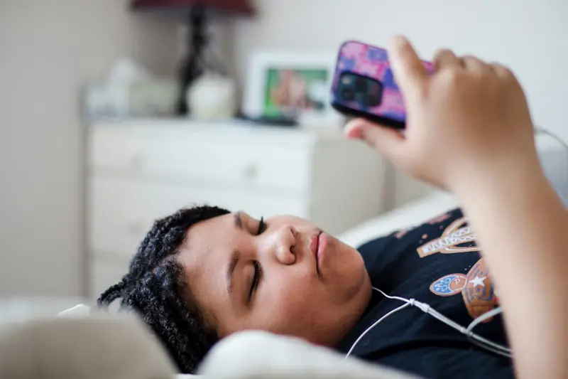 Ein Kind liegt mit Kopfhörern auf dem Bett und hält sein Handy über das Gesicht.