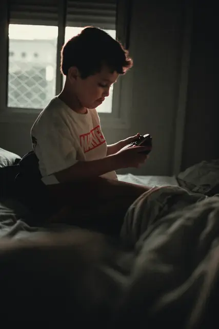 Ein Junge sitzt in einem halbdunklen Zimmer auf dem Bett und spielt auf dem Handy.