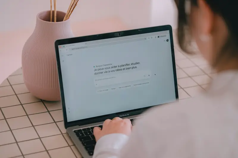 Eine Person tippt am Laptop eine Frage in ein Eingabefeld, daneben steht eine rosa Vase.