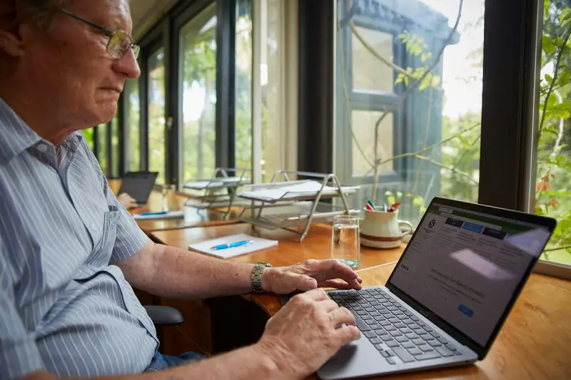 Ein älterer Mann sitzt am Fenster und arbeitet an einem Laptop.
