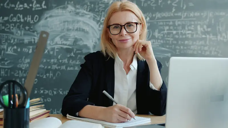 Eine Lehrerin sitzt vor einer vollgeschriebenen Wandtafel am Pult mit Laptop.