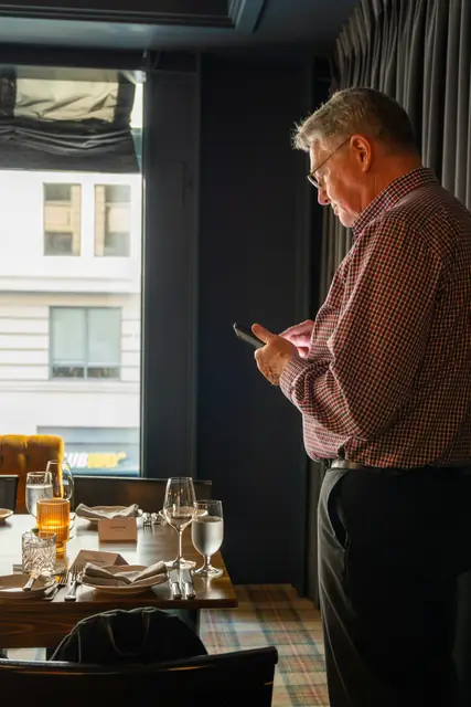 Ein älterer Mann steht in einem Restaurant und liest auf seinem Smartphone.