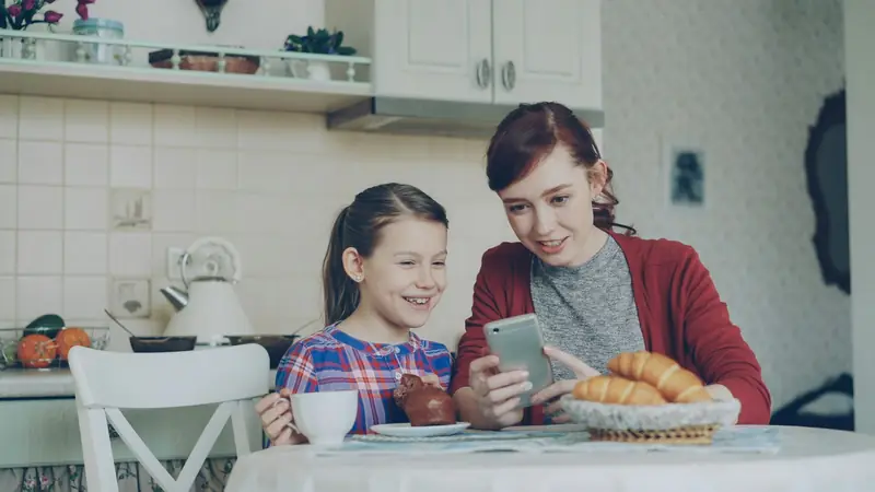 Eine Mutter und ihre Tochter schauen am Küchentisch gemeinsam auf ein Handy.