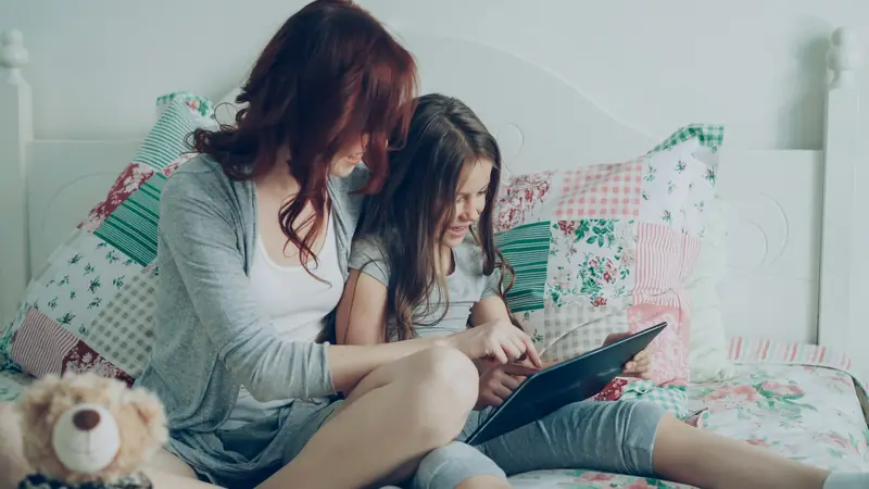 Eine Mutter und ihre Tochter sitzen auf dem Bett und lesen gemeinsam auf einem Tablet.