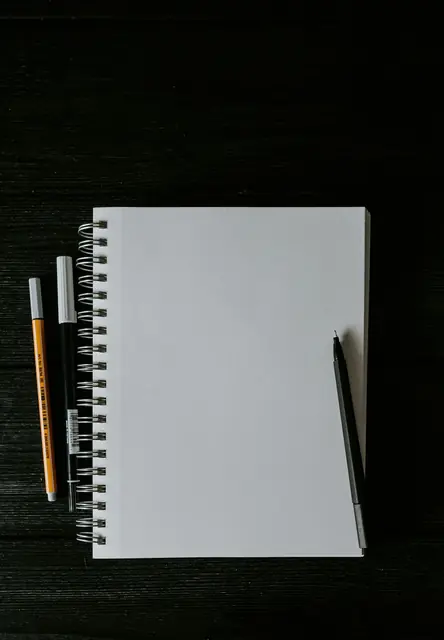 Ein leeres Spiralnotizbuch mit Stiften auf einem dunklen Tisch.