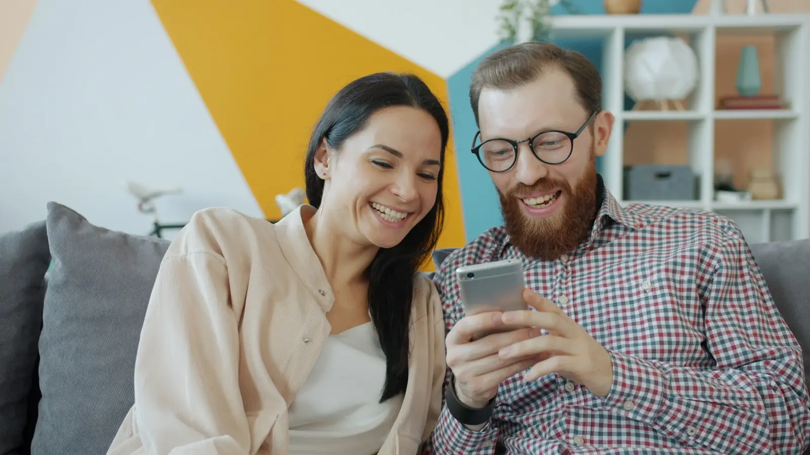 Ein Paar sitzt auf dem Sofa und schaut lachend gemeinsam auf ein Smartphone.