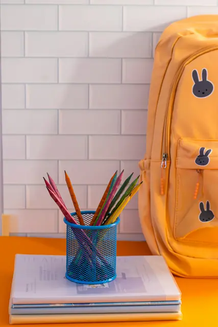 Ein gelber Kinderrucksack und ein Becher voller Farbstifte auf einem Tisch.
