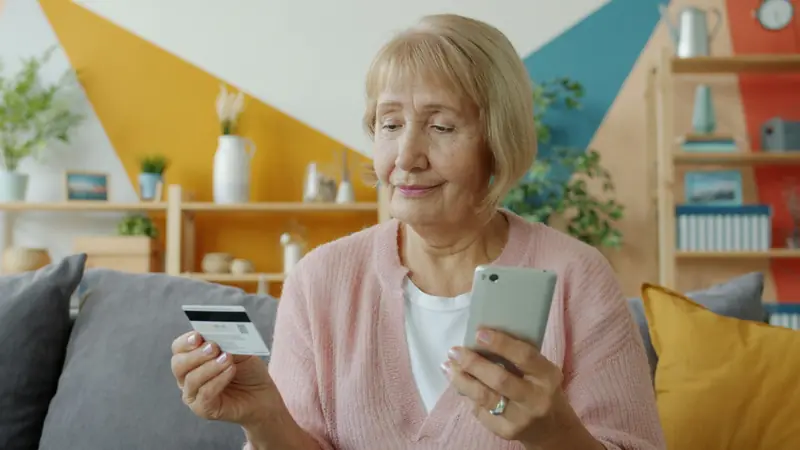Eine ältere Frau hält eine Kreditkarte und schaut prüfend auf ihr Smartphone.
