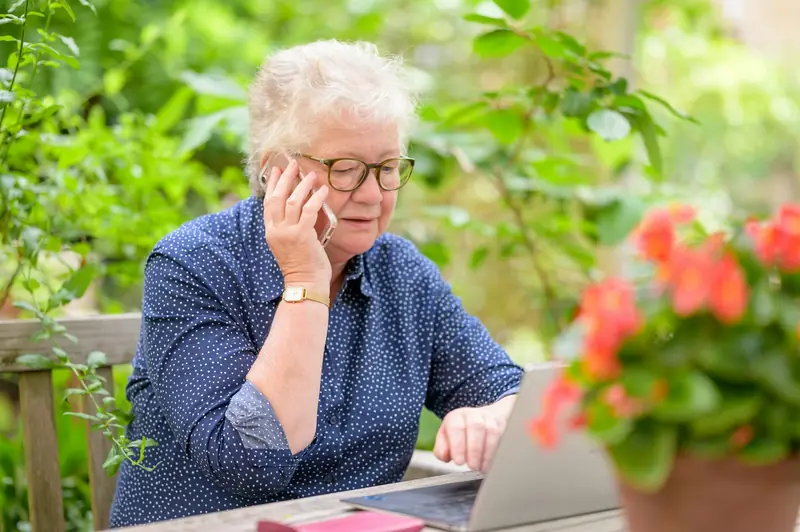 Eine ältere Frau sitzt im Garten vor einem Laptop und telefoniert nachdenklich.